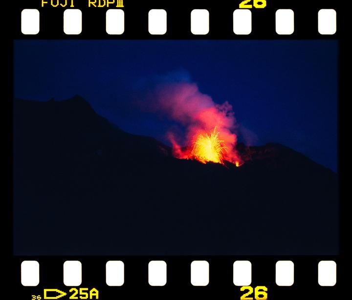 A volcano erupts with bright orange and yellow lava and smoke against a dark blue night sky.