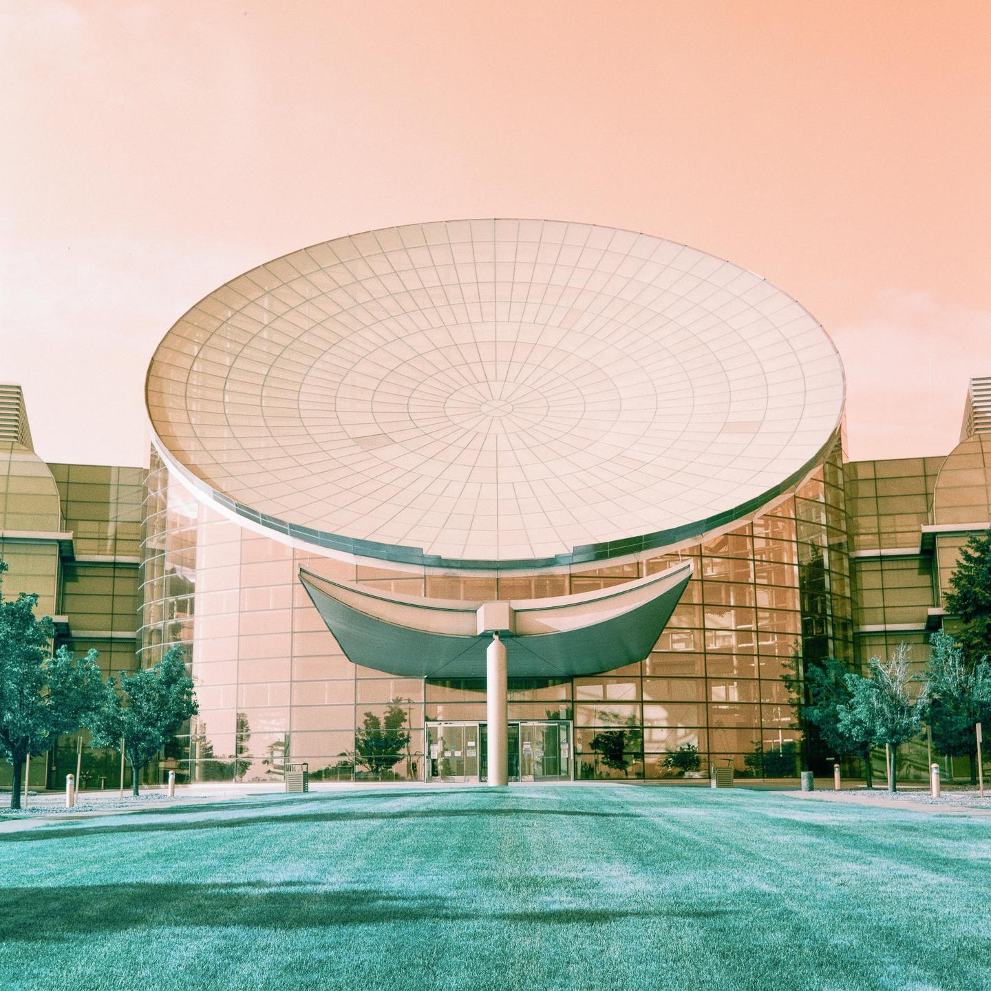 A modern building with a large circular roof and glass facade stands behind a manicured lawn and trees.