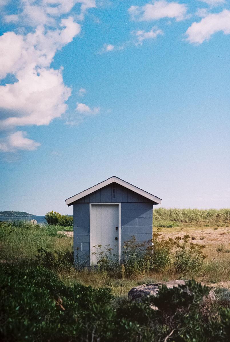 A small blue shack with a white door stands in a grassy field under a blue sky with white clouds.