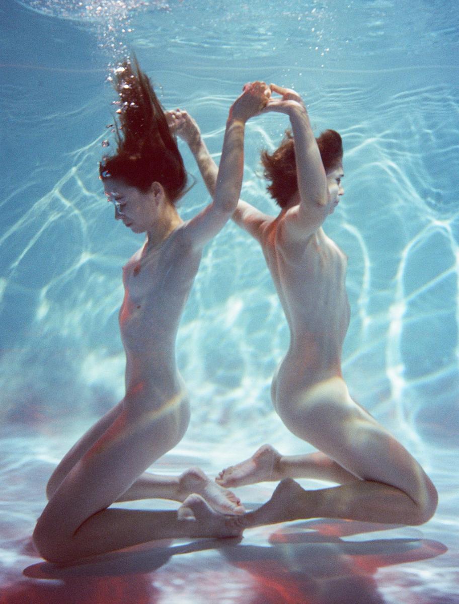 Two women kneel underwater with their arms raised and hands clasped together.