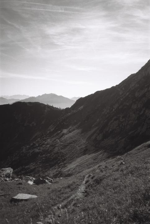 A winding path leads up a grassy mountain slope with distant peaks under a cloudy sky.