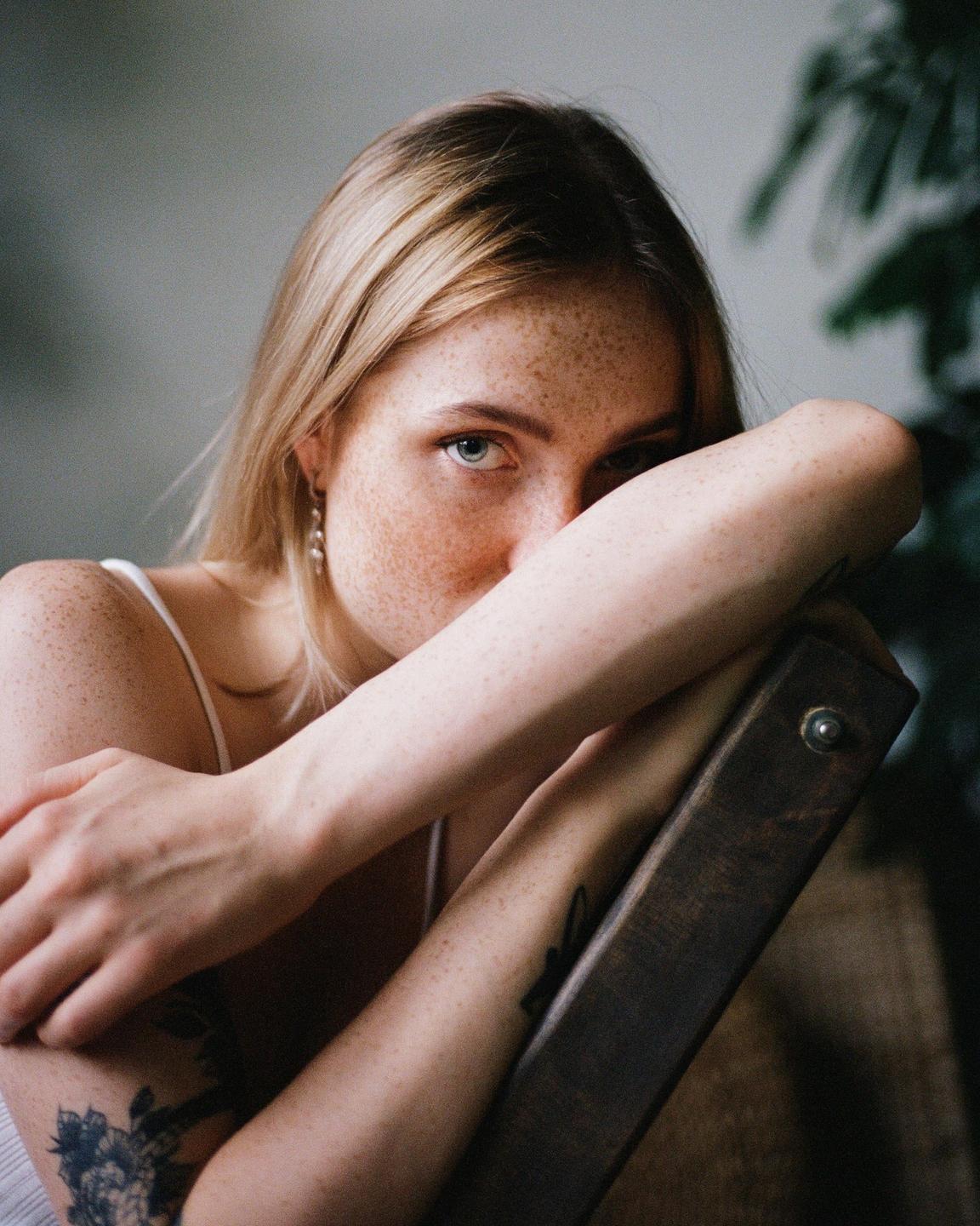 A woman with freckles and blonde hair looks directly at the viewer, resting her arms on a wooden chair.