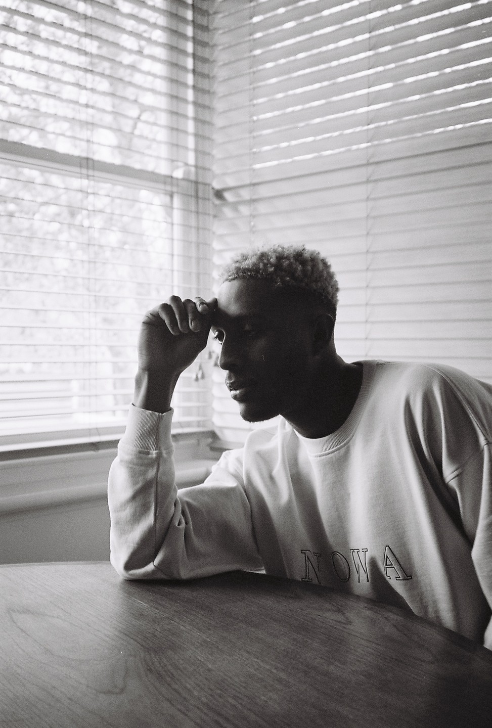 A man with bleached hair sits at a table with his hand on his forehead, looking out a window with venetian blinds.