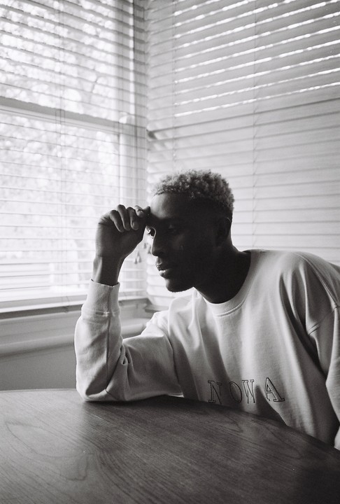 A man with bleached hair sits at a table with his hand on his forehead, looking out a window with venetian blinds.