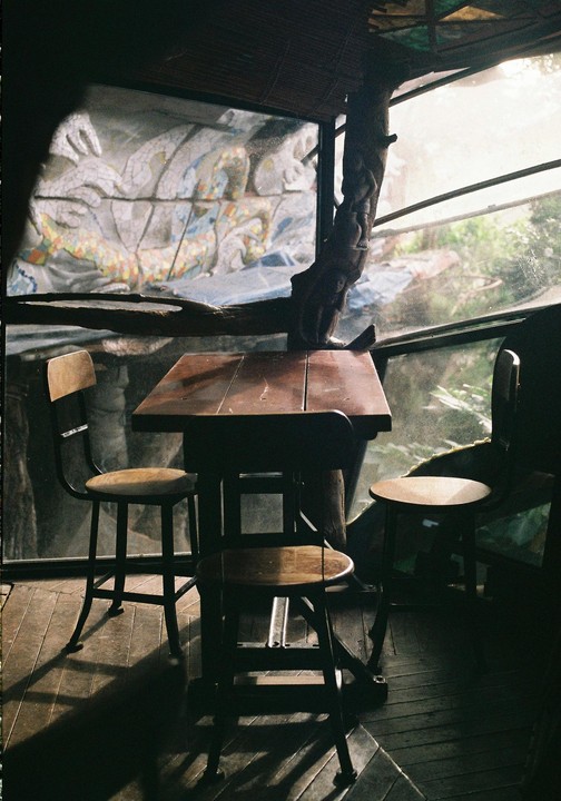 A wooden table and two chairs sit in a dimly lit room with a large window looking out onto a mosaic wall and lush greenery.