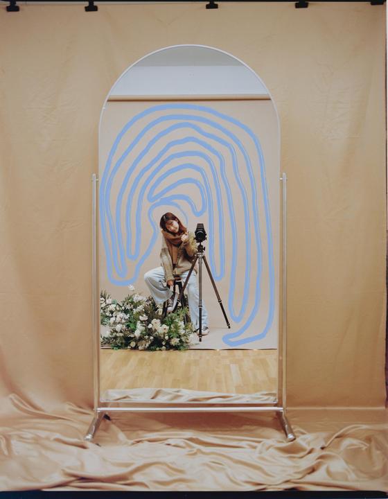 A woman sits on a stool next to a camera on a tripod, surrounded by flowers, reflected in a mirror with blue painted lines.