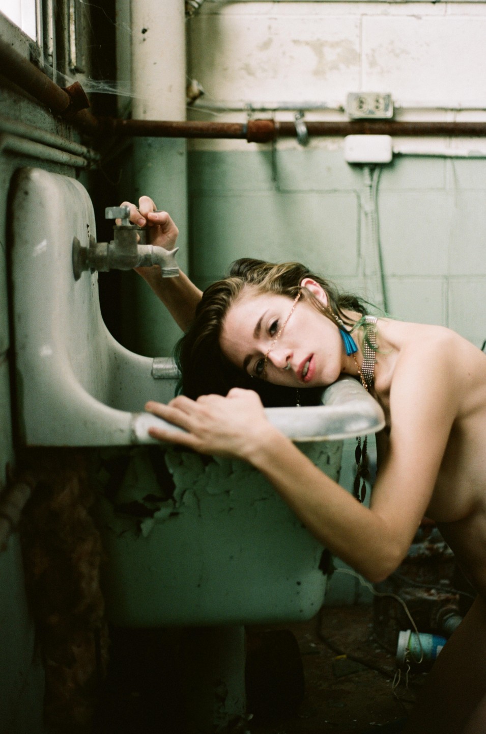 A woman with a chain across her face and a blue earring leans over a dirty sink in an abandoned building.
