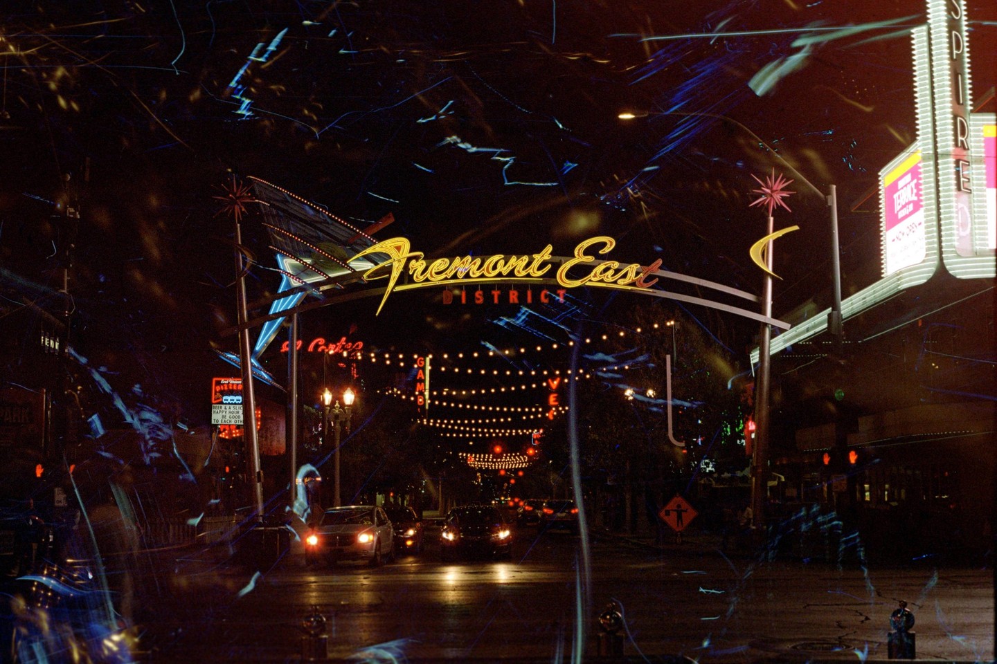 A street at night is illuminated by neon signs and string lights, with cars driving towards the viewer.