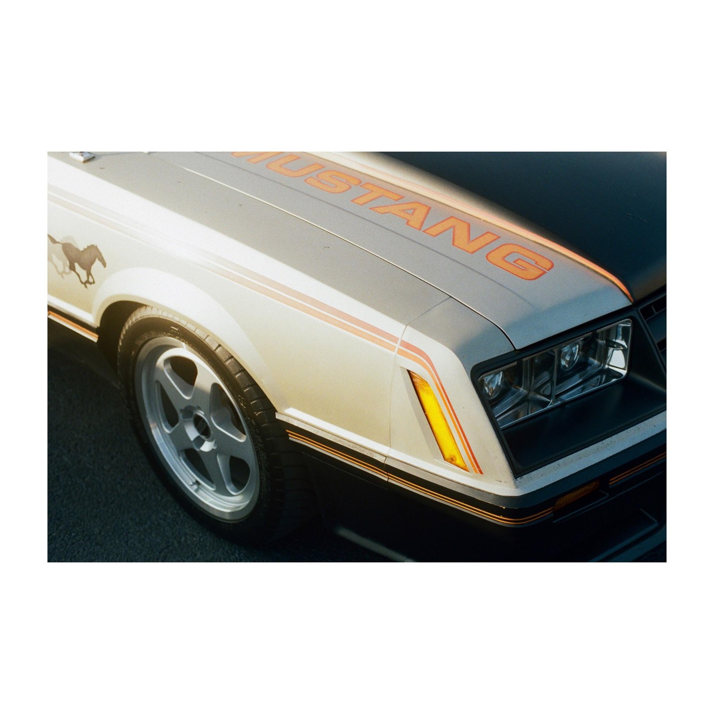 The front quarter panel of a white Ford Mustang is visible, with the word "MUSTANG" in orange letters on the hood.