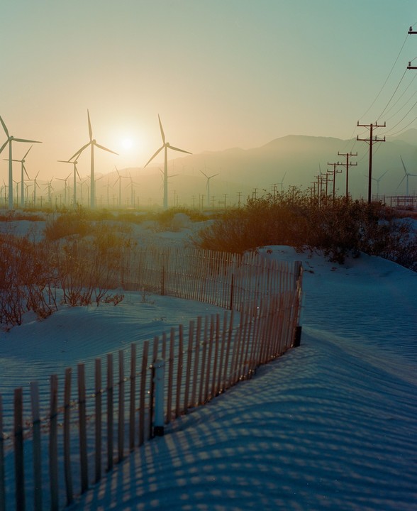 A wooden fence casts shadows on a snowy landscape with wind turbines and mountains in the background during golden hour.