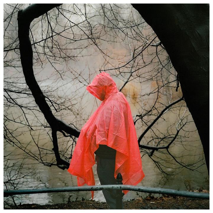 A person in a bright orange poncho stands by a body of water with bare tree branches overhead.