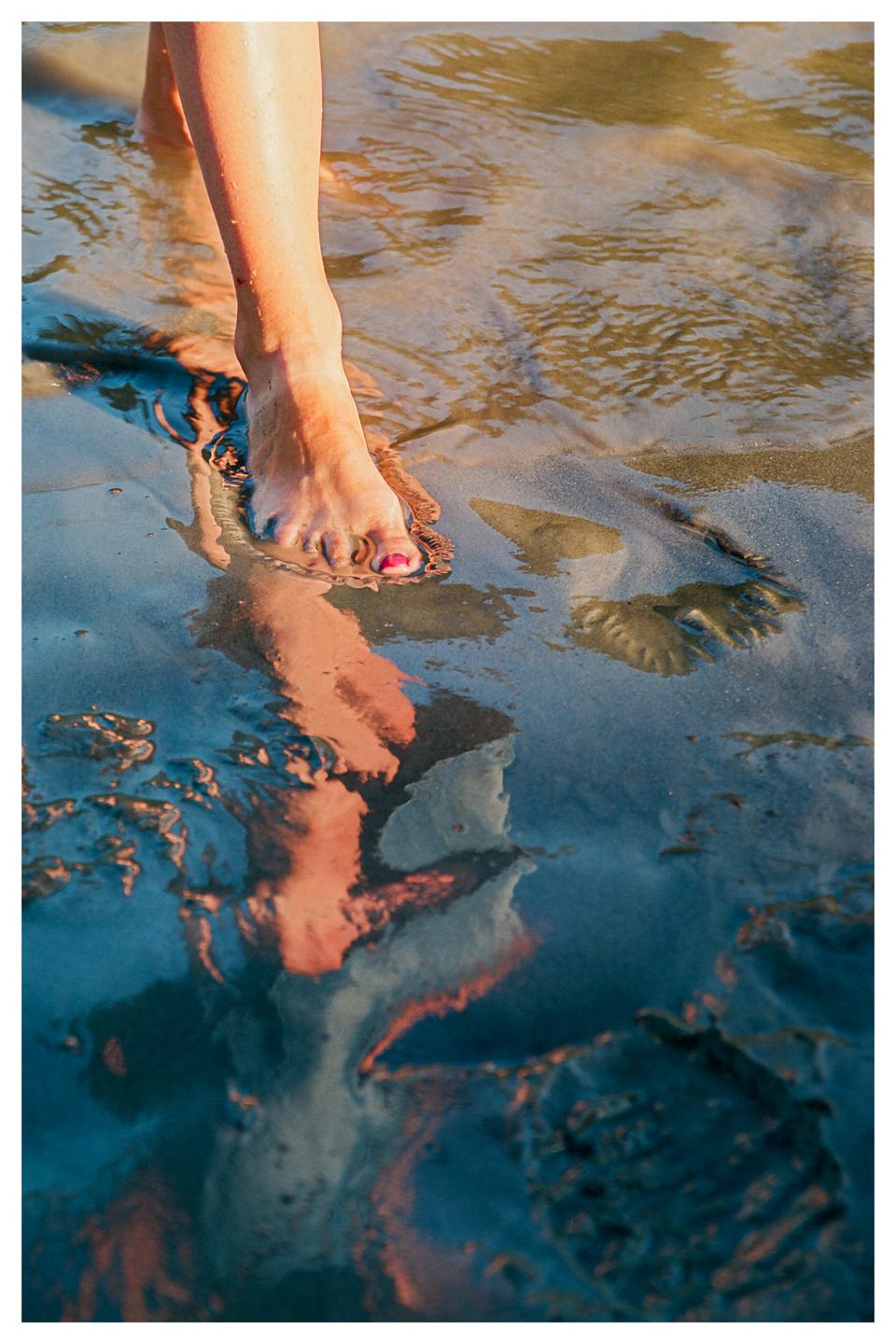 A woman's foot with painted toenails steps into shallow water on a beach during golden hour.