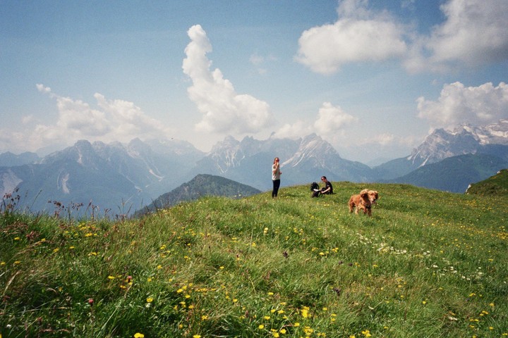 Two women and a dog are on a grassy mountain with a mountain range in the background.