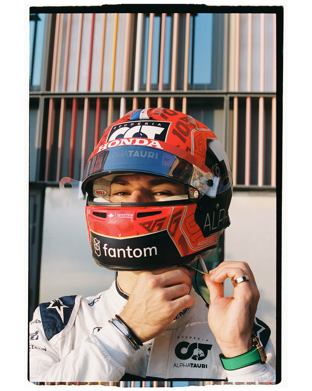 A man in a racing suit adjusts his helmet, which has a red and white design with various logos, against a background of vertical panels.