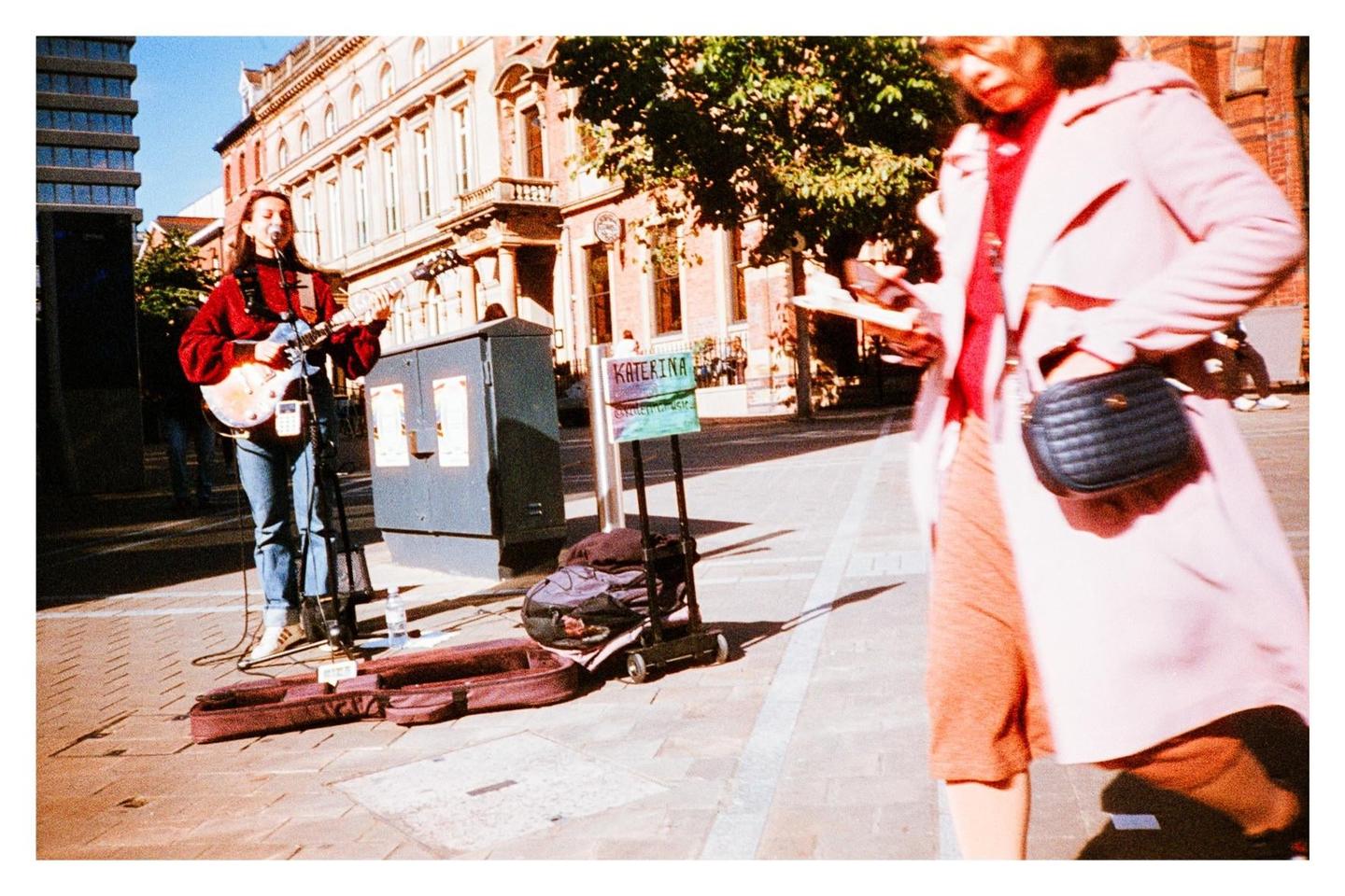 A woman plays a guitar on a city street while another woman walks past in the foreground.