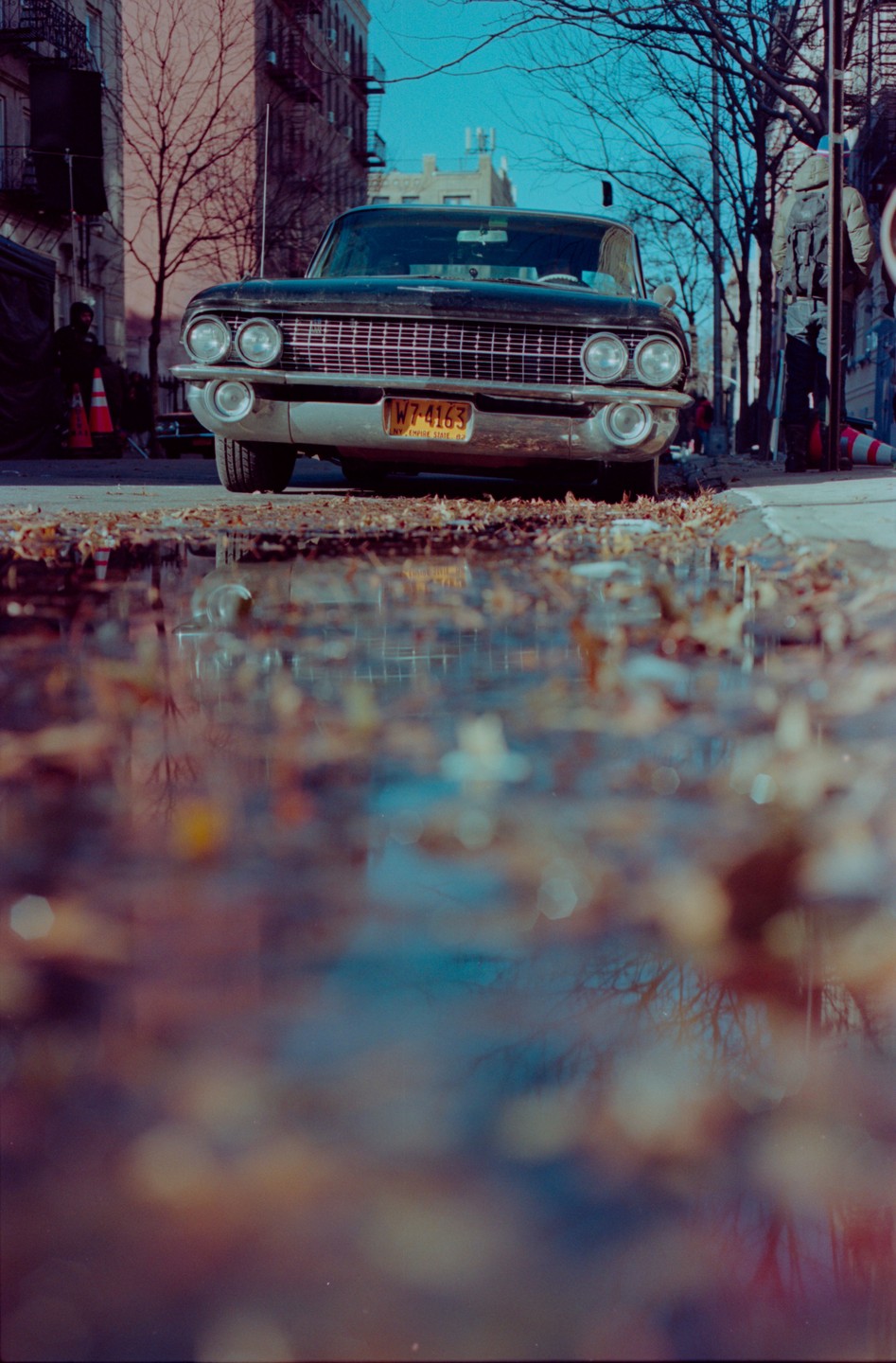 A black car is parked on a street next to a puddle reflecting the sky and trees.
