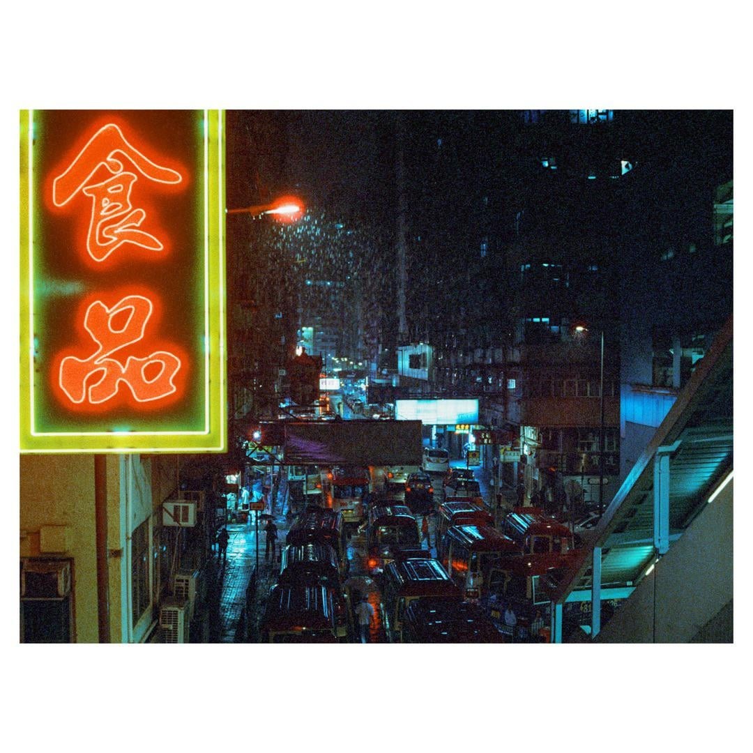 A neon sign illuminates a busy street filled with vehicles and pedestrians on a rainy night.