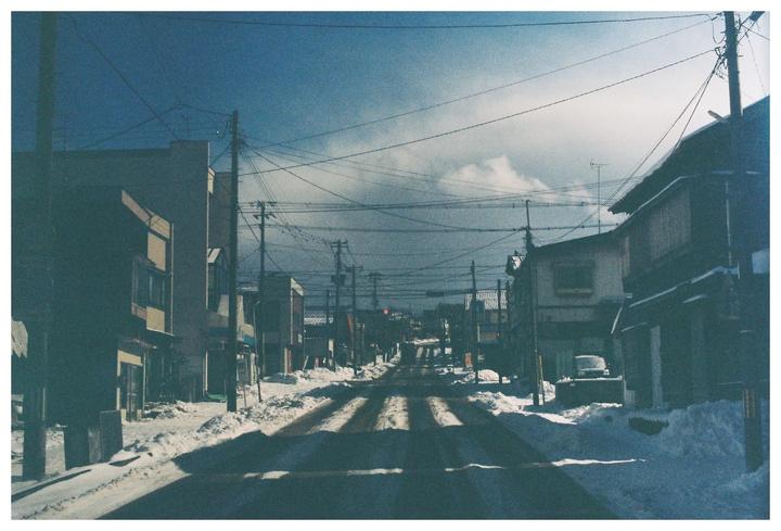 A snow-covered street with buildings and utility poles on both sides stretches into the distance under a cloudy sky.