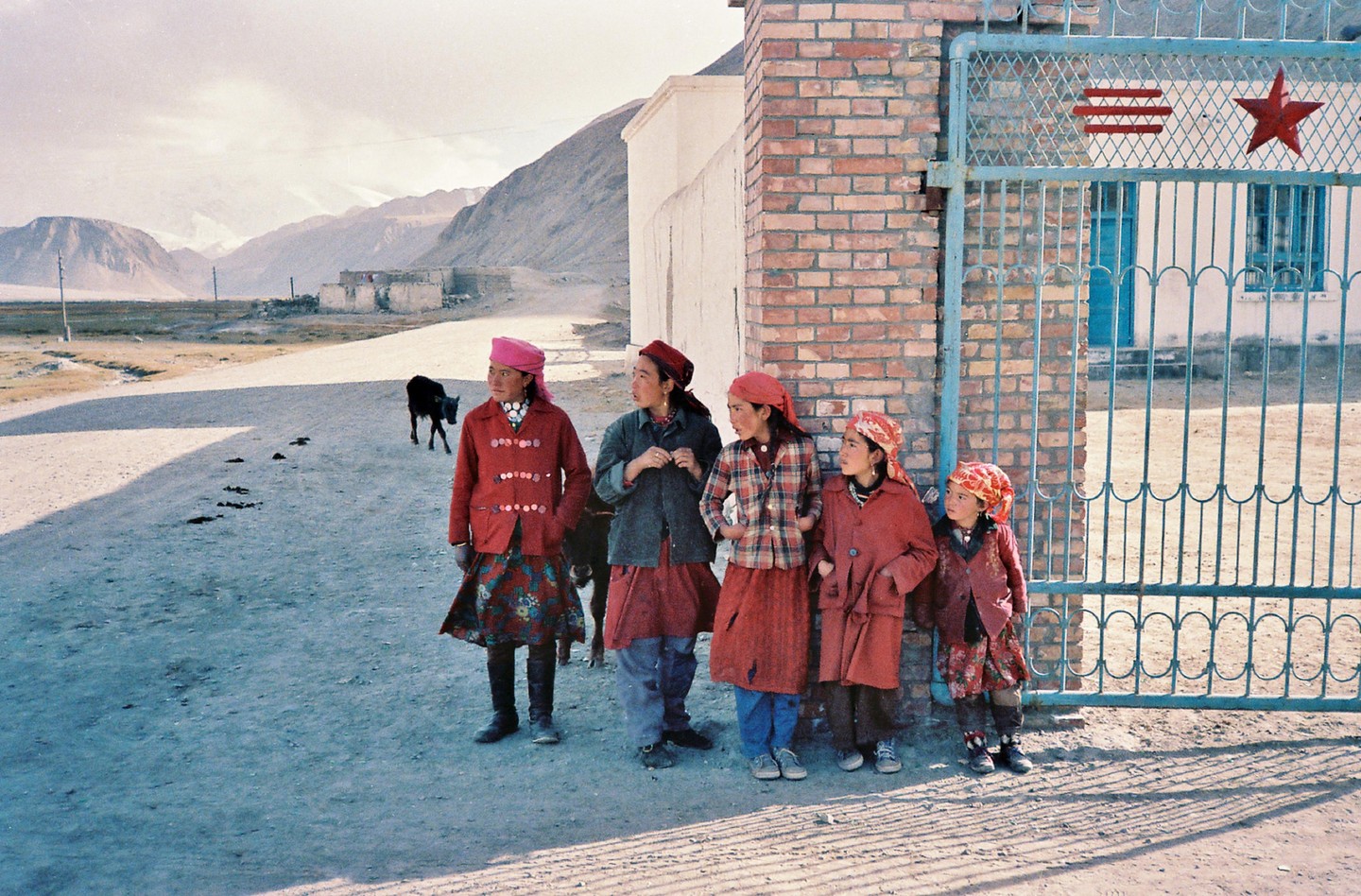 Five girls stand by a brick wall and a blue metal gate with a red star, with mountains and a dirt road in the background.