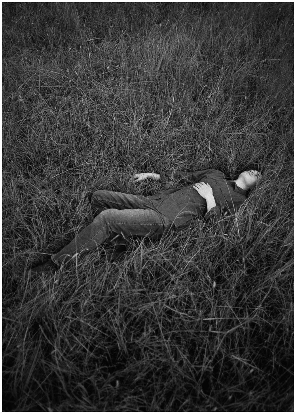 A man lies in tall grass with his eyes closed and one hand on his chest.