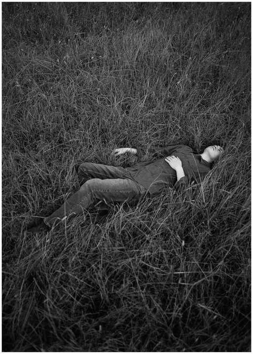 A man lies in tall grass with his eyes closed and one hand on his chest.