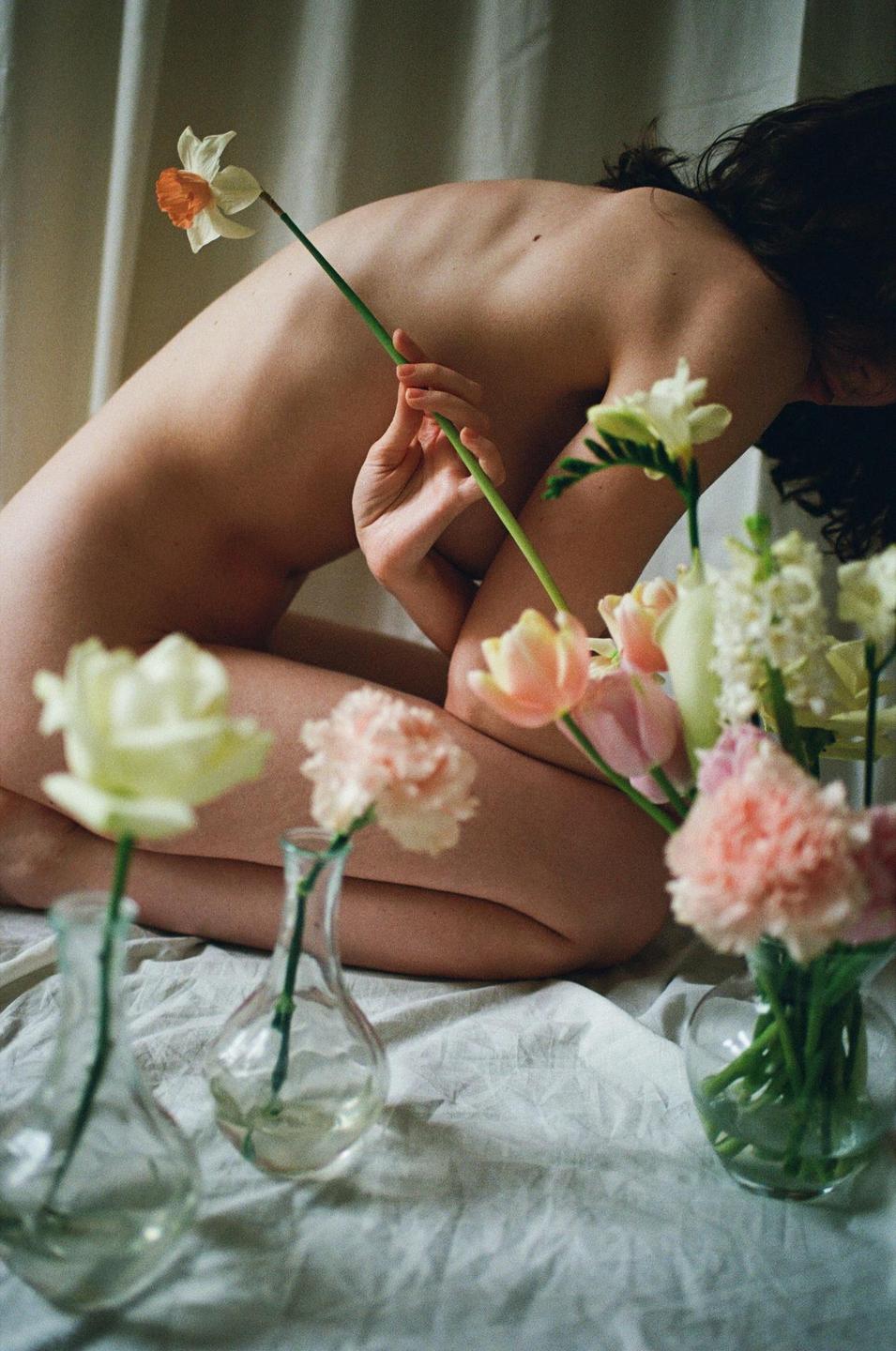 A woman kneels on a white sheet, holding a daffodil, surrounded by flowers in vases.
