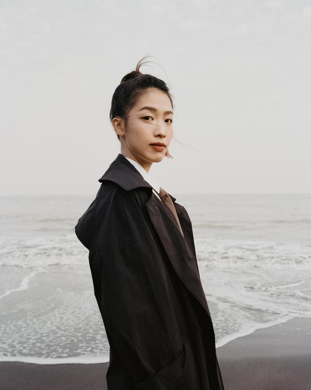 A woman in a dark coat stands on a beach with waves crashing behind her.