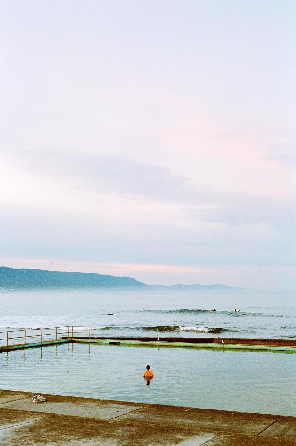 A person in an orange shirt stands in a concrete ocean pool with mountains and surfers in the background.