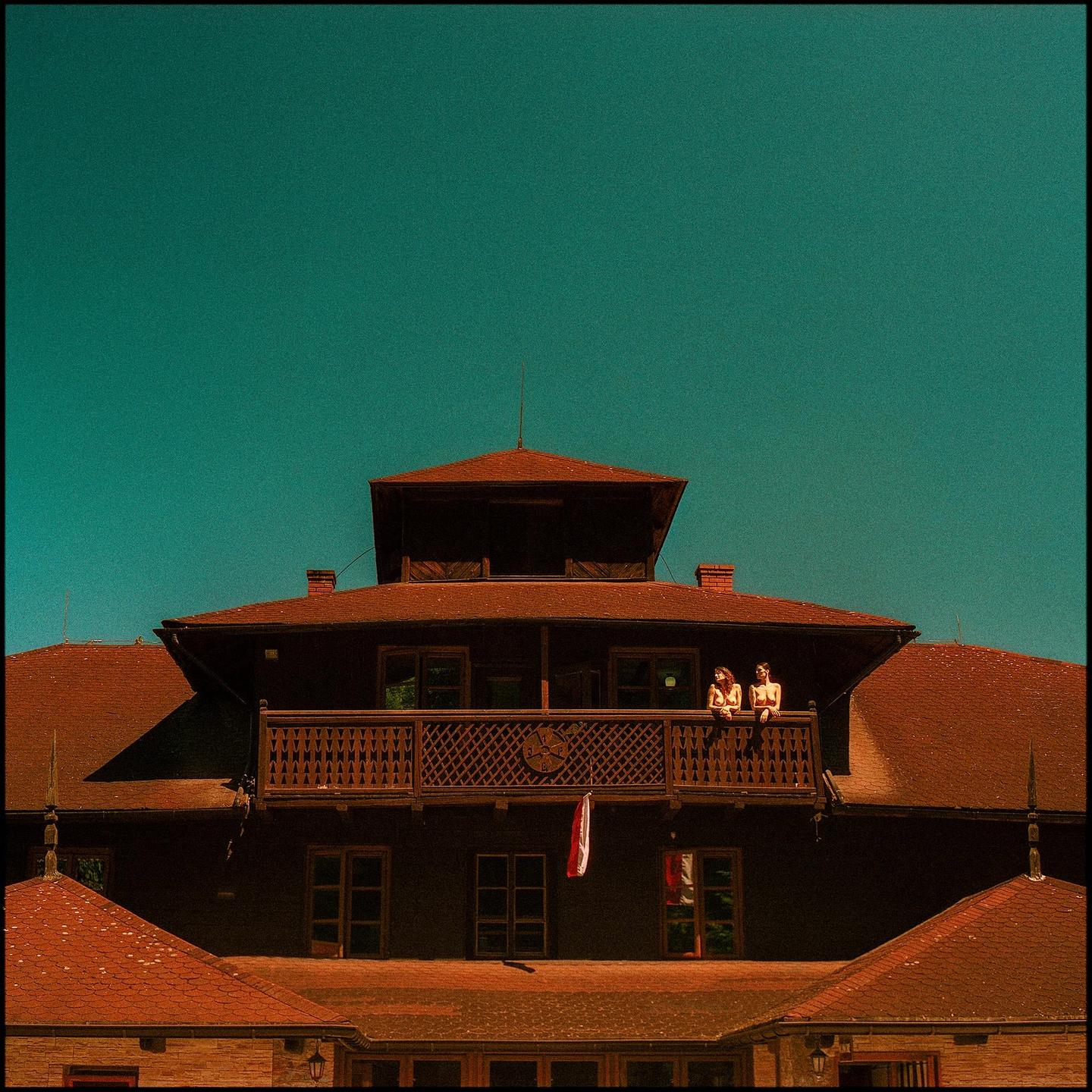 Two women stand on a balcony of a guesthouse with a red roof under a clear blue sky.
