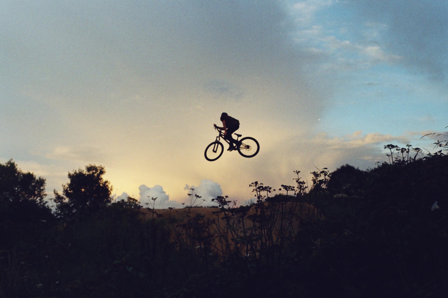 A silhouetted person on a bicycle is airborne against a sunset sky with clouds and dark foliage below.