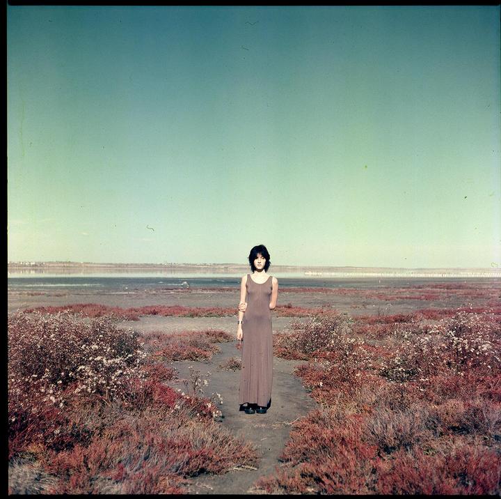 A woman stands in a barren landscape with red bushes and a body of water in the distance.