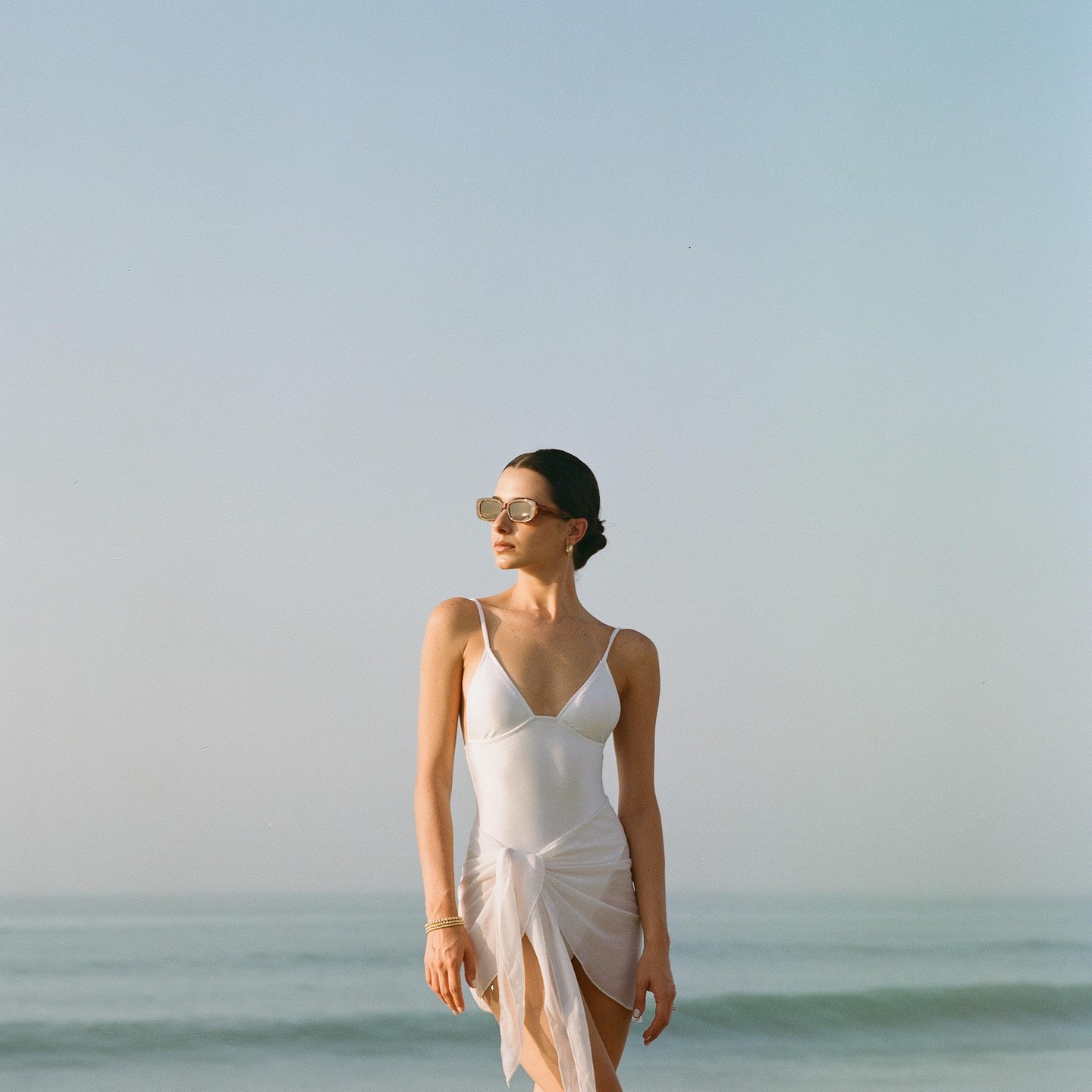 A woman in a white swimsuit and sunglasses stands on a beach looking out at the ocean.