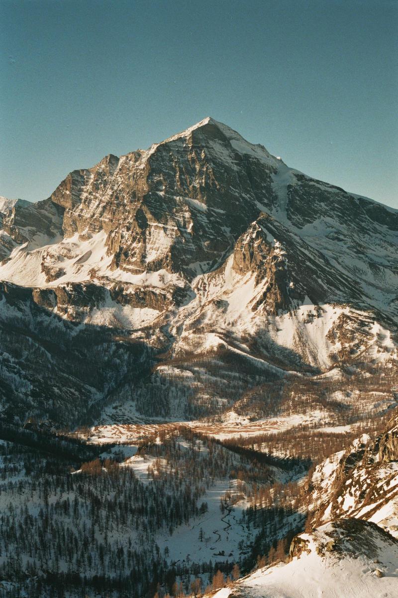 A snow-covered mountain peak rises above a valley with scattered trees and patches of snow.