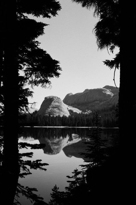 A lake reflects a large rock formation and a forest under a bright sky, framed by dark trees.