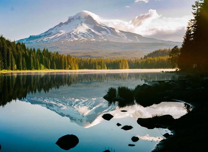A snow-capped mountain with a cloud on its peak is reflected in a still lake surrounded by a forest.