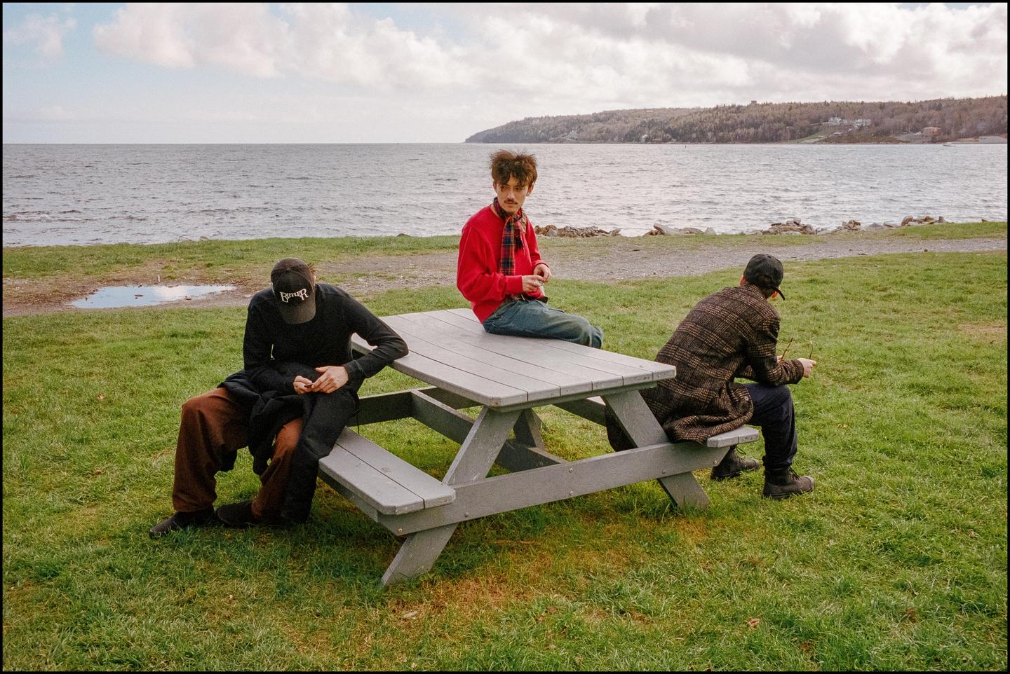 Three men are sitting on and around a picnic table by the water.