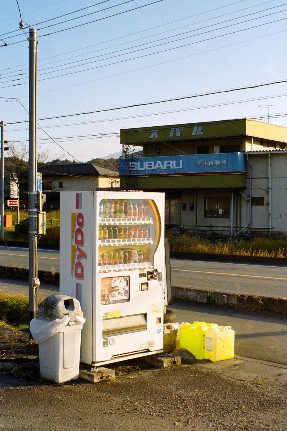 A DyDo vending machine stands next to a trash can on the side of a road, with a Subaru building in the background.