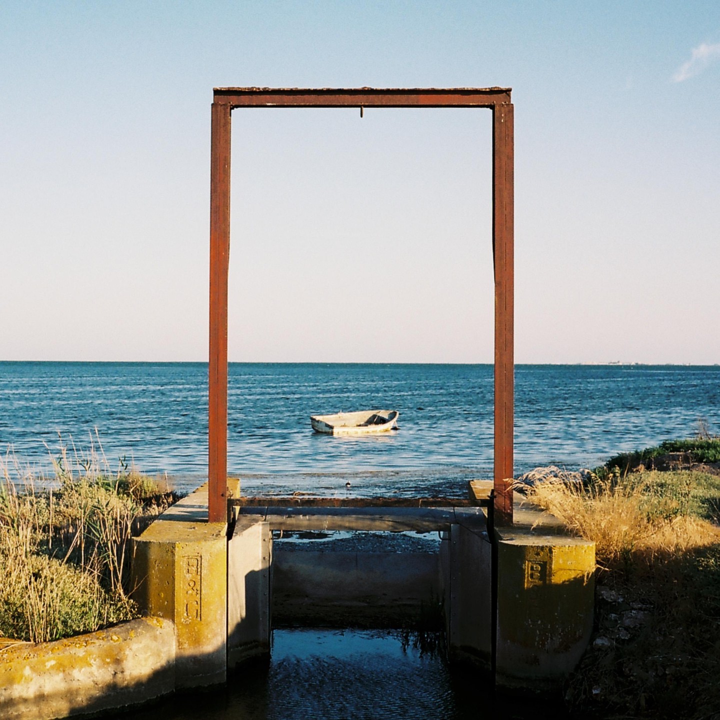 A small boat floats in the sea, framed by a large metal structure on the shore.