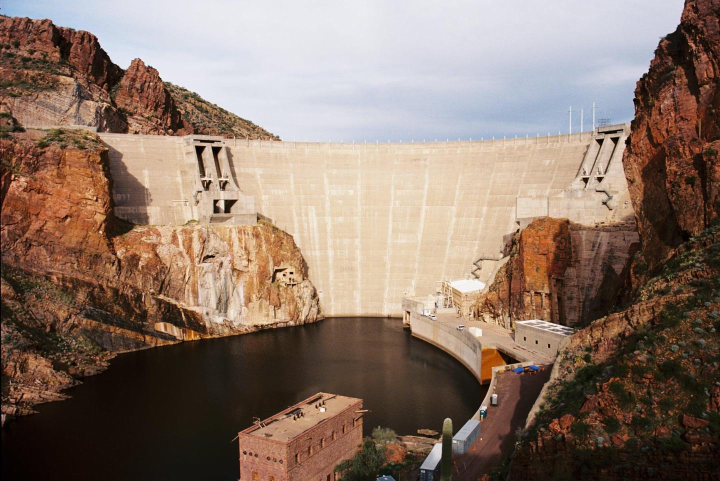 A large concrete dam is nestled between two rocky mountains, with a body of water at its base and a building on the right.
