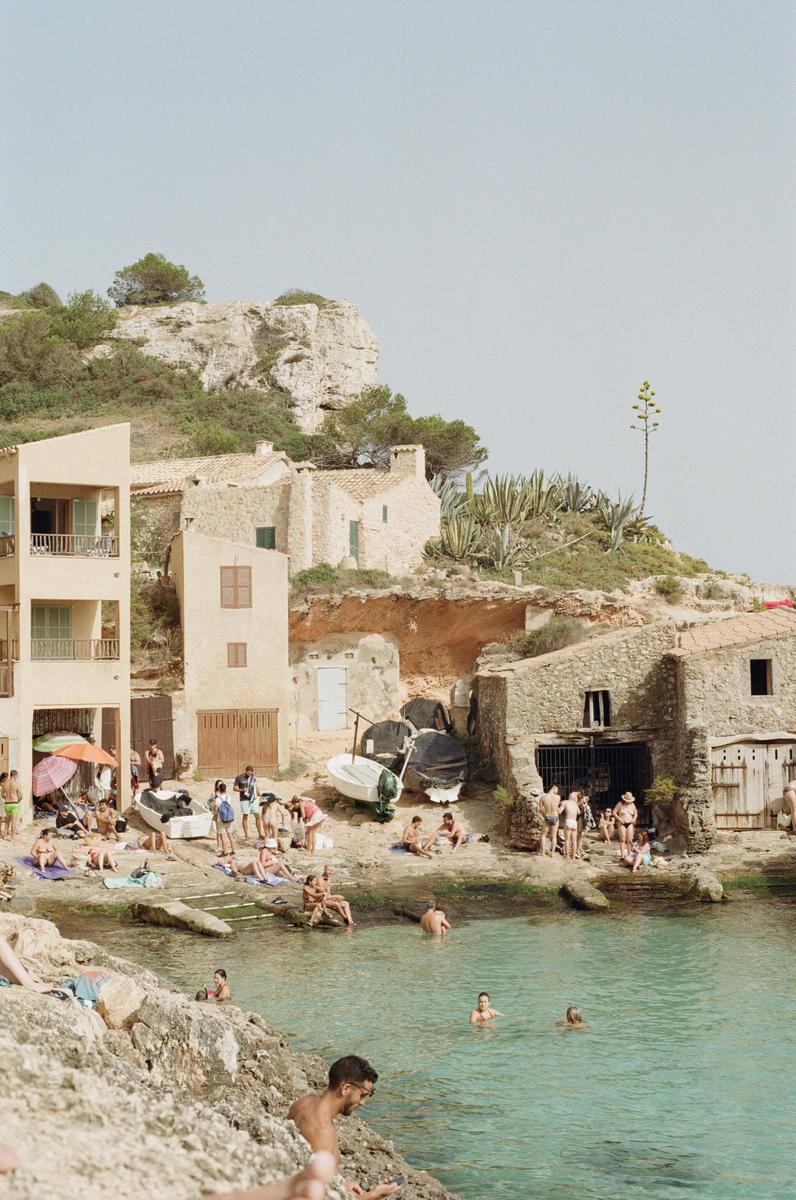 A group of people are swimming and relaxing on a rocky beach with buildings and cliffs in the background.
