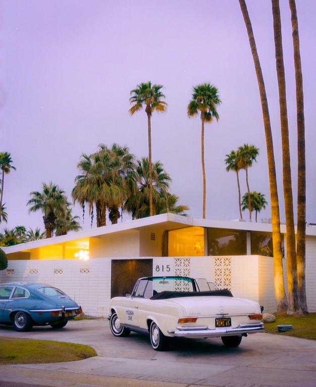 Two vintage cars are parked in front of a mid-century modern house with palm trees under a purple sky.
