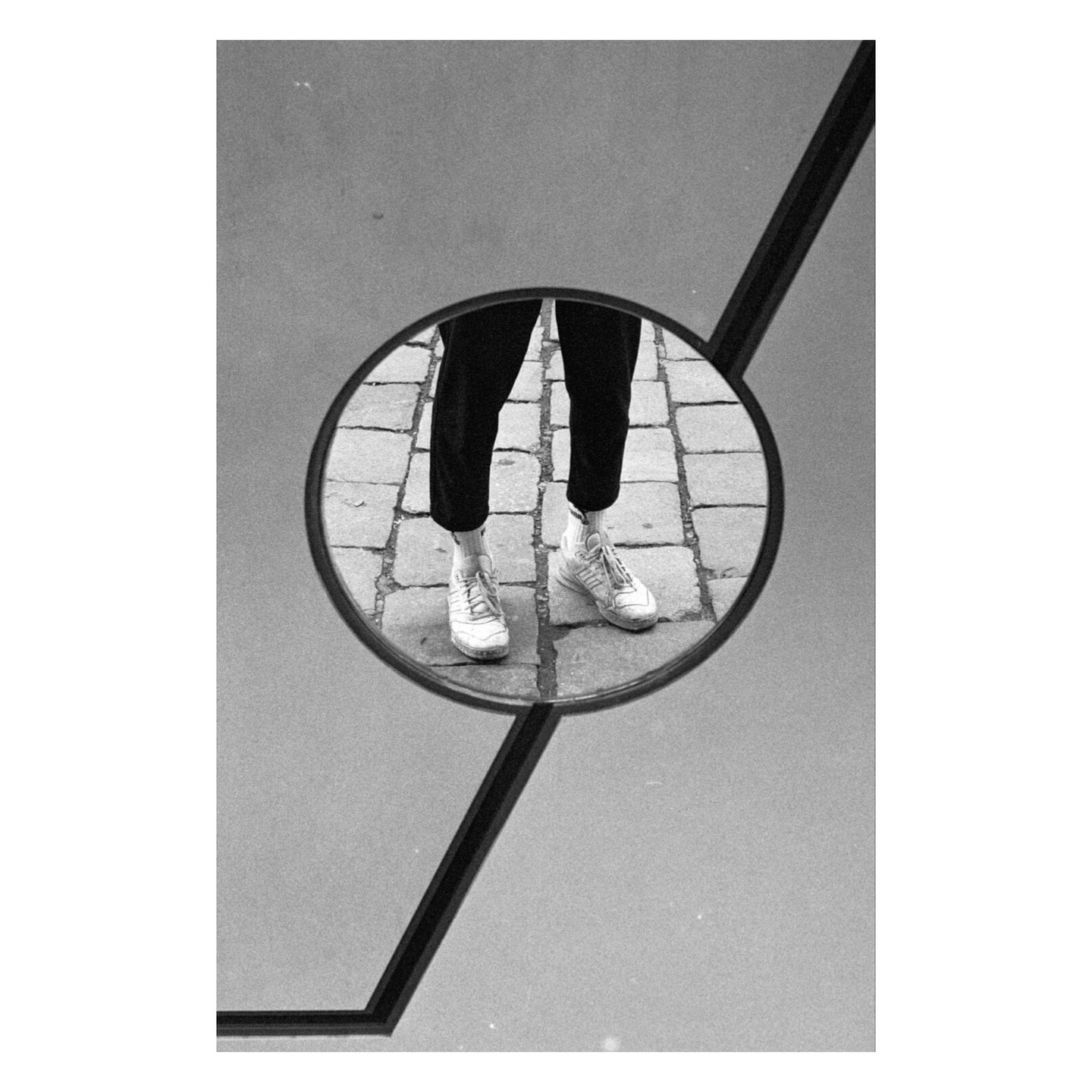 A person's legs and feet are reflected in a circular mirror, showing white sneakers on a cobblestone street.