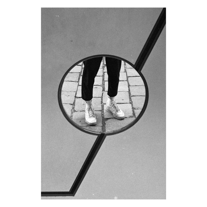 A person's legs and feet are reflected in a circular mirror, showing white sneakers on a cobblestone street.