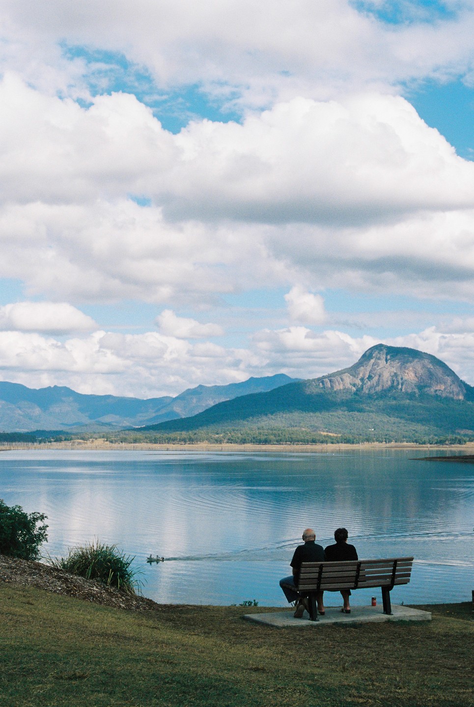 A couple sits on a bench overlooking a lake with mountains in the background.