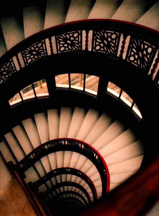 A spiral staircase with ornate railings descends into darkness, with windows visible on the upper landing.