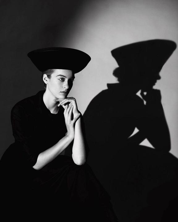 A woman in a hat sits with her hands clasped under her chin, casting a large shadow on the wall behind her.