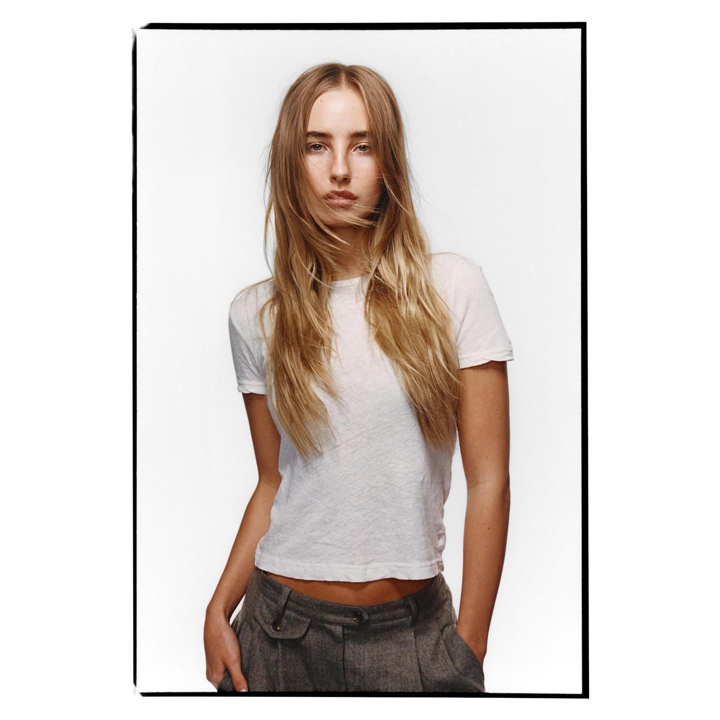 A woman with long blonde hair and a white t-shirt stands with her hands in her pockets.