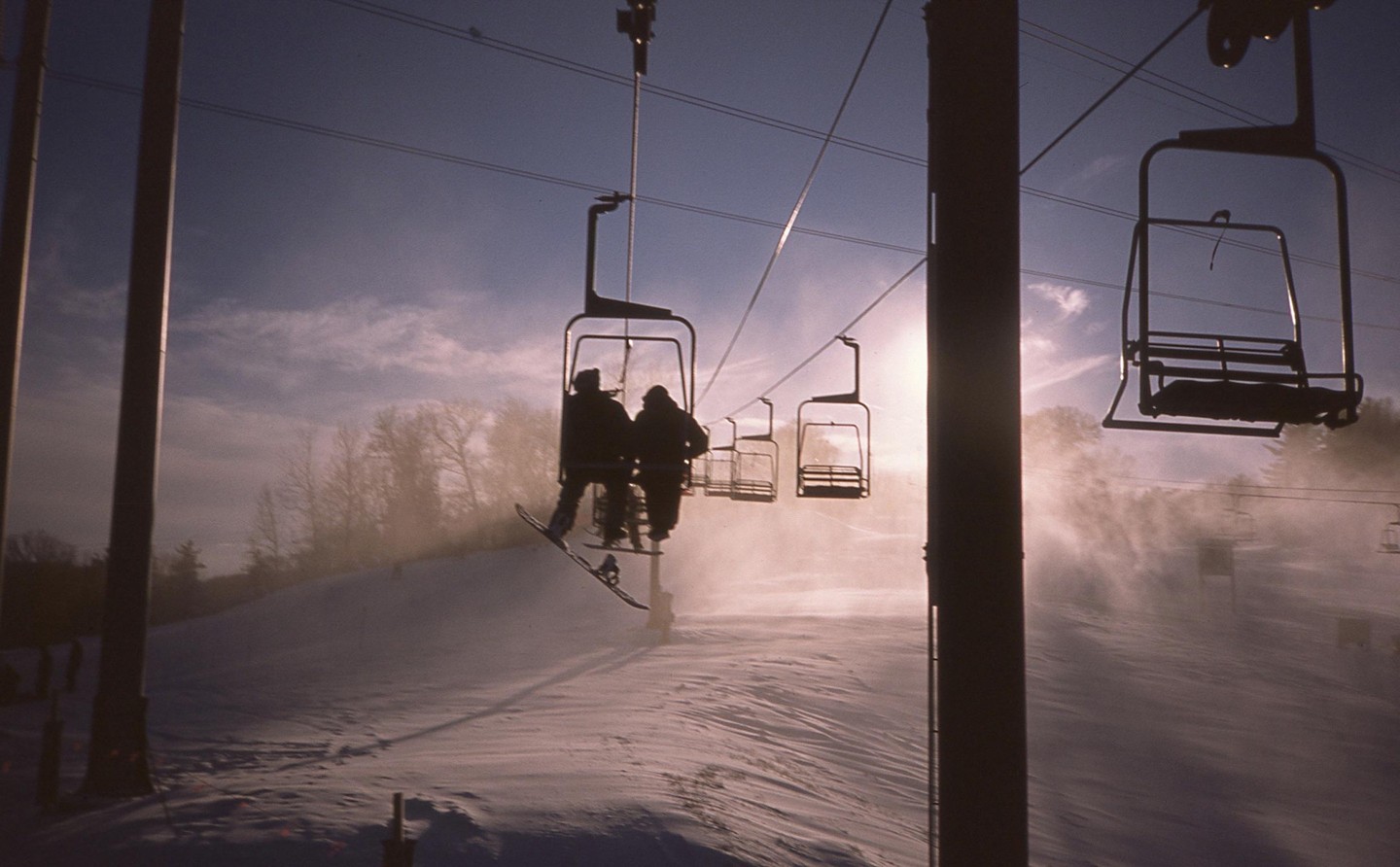 Two people ride a ski lift up a snowy mountain with the sun shining brightly behind them.