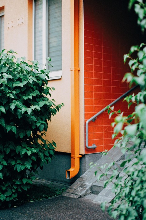 A bright orange downspout runs down the side of a building with a red tiled wall and a green bush in the foreground.