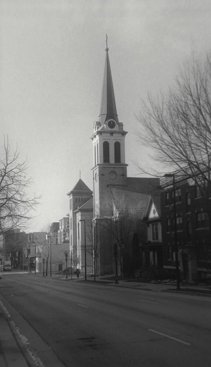 A tall church steeple with a clock is visible from a street lined with bare trees and buildings.